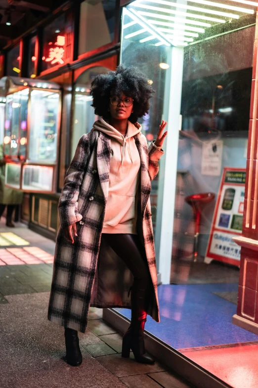 a woman standing on a sidewalk in front of a store, stylish coat for a rave, flannel, with afro, night setting