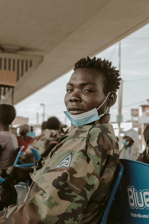 a man sitting in a chair wearing a face mask, by Sebastian Vrancx, trending on unsplash, afrofuturism, wearing camo, in front of a large crowd, in africa, teenage boy