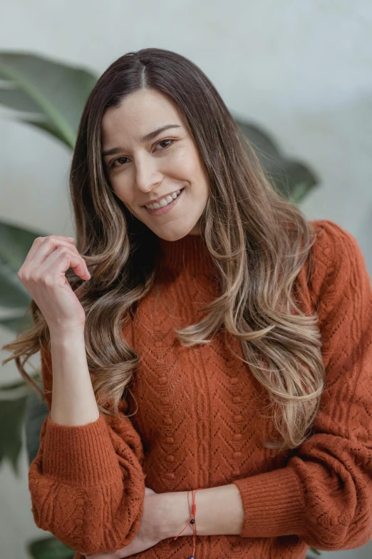 a woman standing in front of a potted plant, by Robbie Trevino, trending on pexels, renaissance, he is wearing a brown sweater, long brown wavy hair, elegant smiling pose, ombre