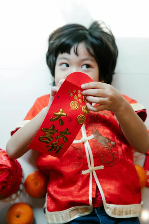 a little girl holding a red packet in front of a pile of oranges, inspired by Mi Fu, pexels, happening, ornate mask and fabrics, wearing a red outfit, cute boy, opening