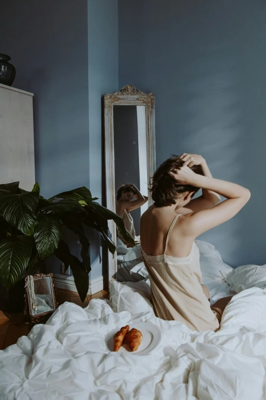 a woman sitting on a bed in front of a mirror, inspired by Elsa Bleda, trending on pexels, her hair is tied above her head, deep blue mood, covered in plants, waking up