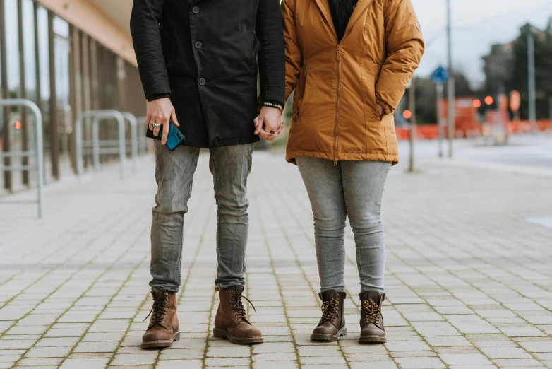 a man and a woman standing next to each other, trending on pexels, antipodeans, brown boots, lgbtq, background image, brown jacket