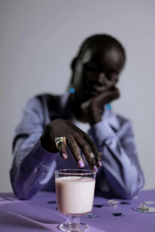 a woman sitting at a table with a glass of milk, by Nathalie Rattner, trending on unsplash, afrofuturism, soft purple glow, photographed for reuters, african man, adut akech