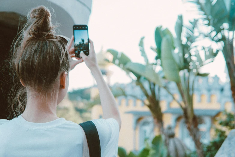 a woman taking a picture with her cell phone, trending on pexels, with palm trees in the back, girl with messy bun hairstyle, from a distance, colour photograph