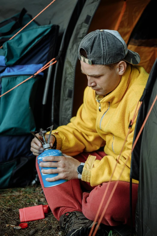 a man sitting on the ground in front of a tent, holding flask in hand, wearing a yellow hoodie, multicoloured, thumbnail