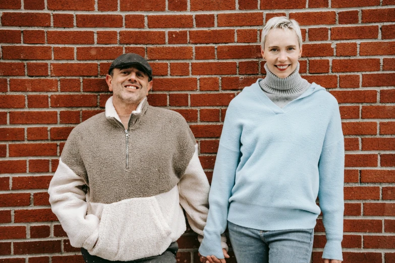 a man and a woman standing in front of a brick wall, by Terese Nielsen, unsplash, wearing casual clothing, warm features, two old people, background image