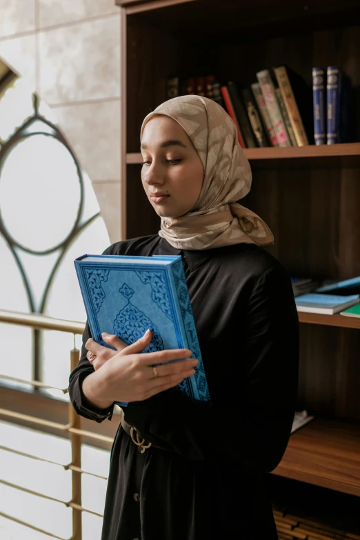 a woman standing in front of a bookshelf holding a book, by Julia Pishtar, trending on pexels, hurufiyya, head scarf, holy, blue, holding notebook