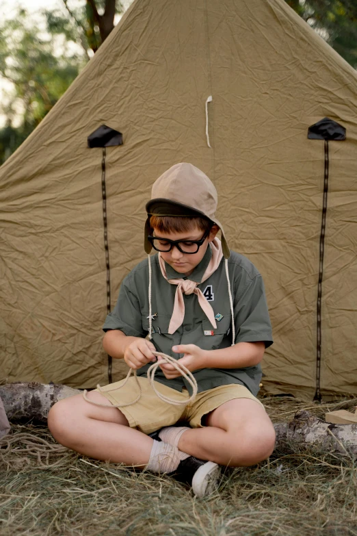 a woman sitting on the ground in front of a tent, inspired by Wes Anderson, trending on pexels, boy scout troop, stitching, caracter with brown hat, cute boy