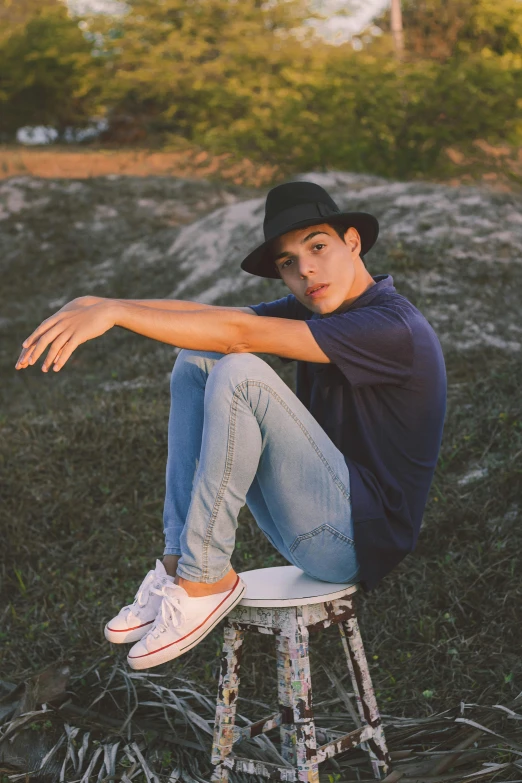 a man sitting on top of a stool in a field, pexels contest winner, realism, teenage jughead jones, confident relaxed pose, with hat, caio santos