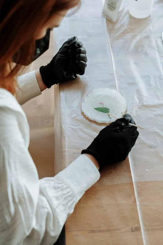 a woman in black gloves sitting at a table, a detailed painting, inspired by Master of the Embroidered Foliage, trending on pexels, process art, wearing translucent sheet, resin and clay art, leaves and simple cloth, white