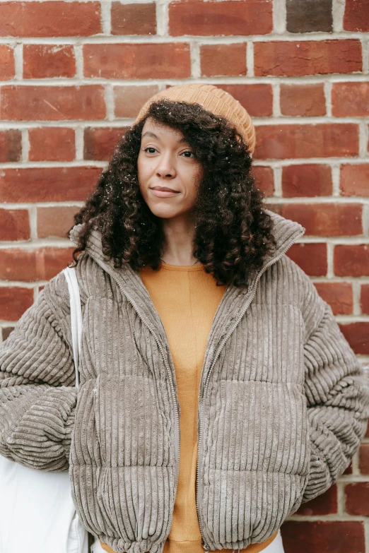 a woman standing in front of a brick wall, trending on pexels, renaissance, puffer jacket, mixed race woman, corduroy, knitted hat