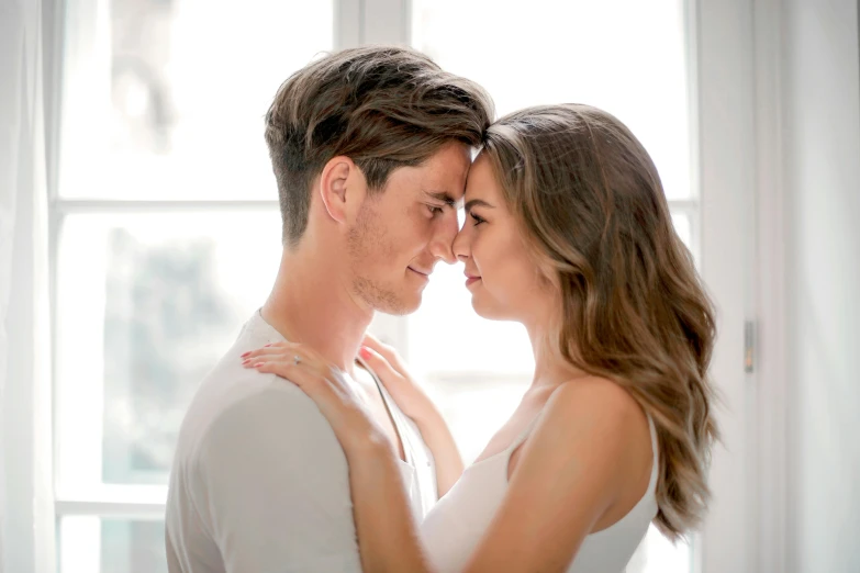 a man and woman embracing in front of a window, trending on pexels, slight stubble, brunette, facing each other, natural light in room