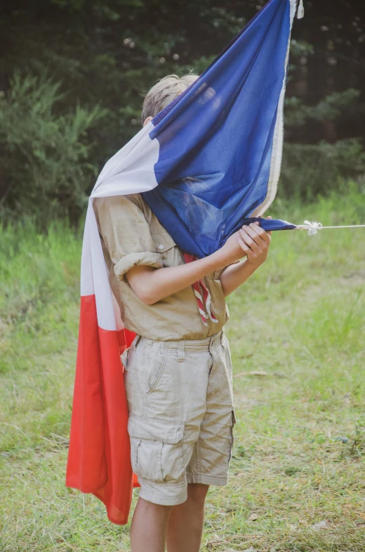 a boy standing in a field holding a flag, by Raphaël Collin, unsplash, renaissance, boy scout troop, french kiss, old color photo, he is at camp