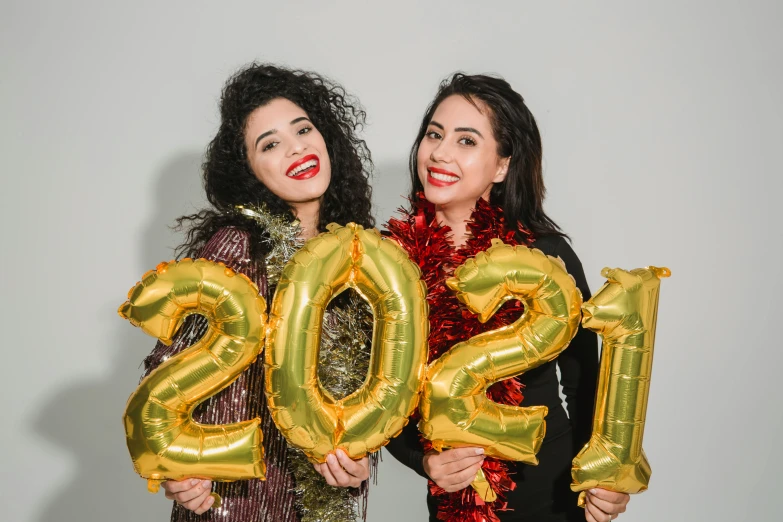 a couple of women standing next to each other holding balloons, trending on pexels, happening, golden number, new years eve, thumbnail, cardboard