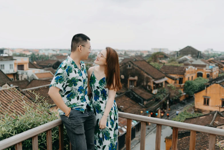 a man and woman standing next to each other on a balcony, inspired by Ruth Jên, pexels contest winner, vietnamese woman, background image, town in the background, wearing a hawaiian dress