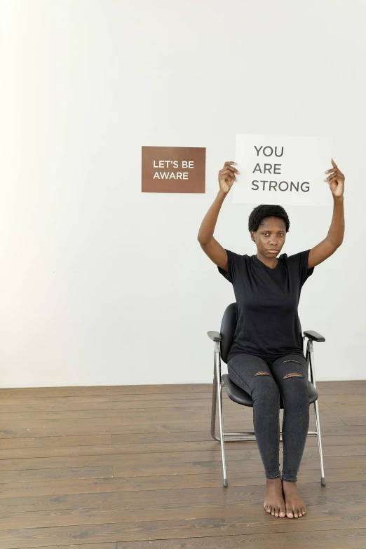 a woman sitting in a chair holding a sign, inspired by Carrie Mae Weems, trending on unsplash, feminist art, strong arms, a person standing in front of a, athletic and strong, you