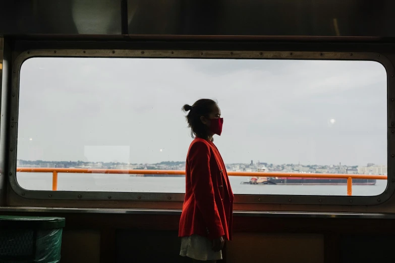 a woman standing in front of a window looking out at a body of water, people are wearing masks, wearing red jacket, interior of staten island ferry, unsplash photography