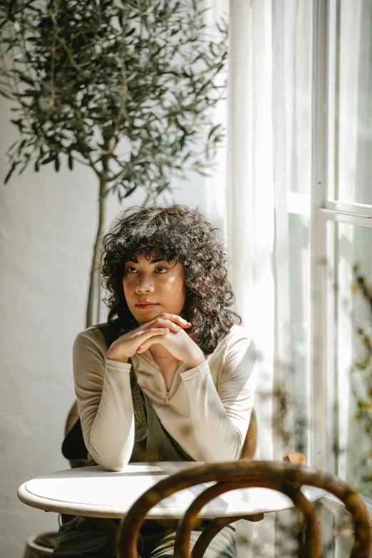 a woman sitting at a table in front of a window, a portrait, pexels contest winner, black curly hair, thinker pose, curly bangs, botanicals