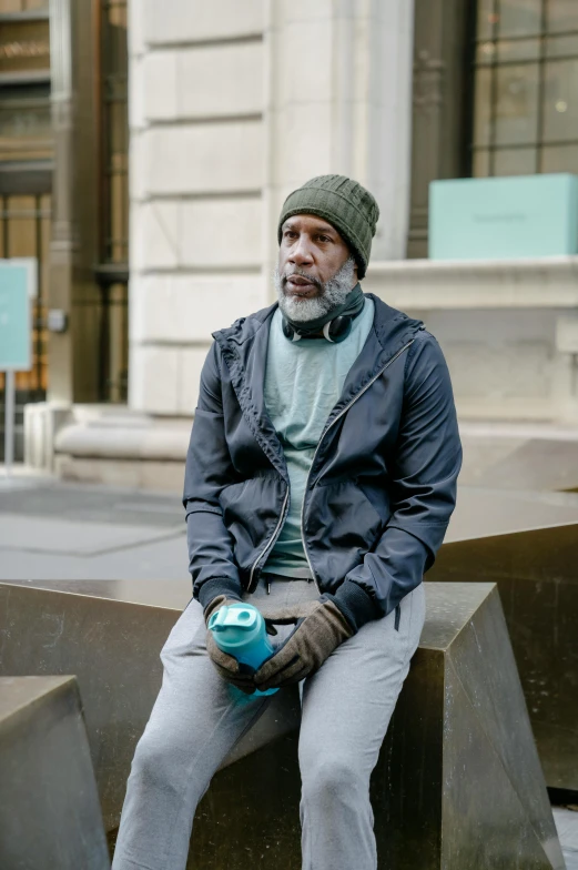 a man sitting on a bench in front of a building, wearing teal beanie, greg irons, in new york, gray beard