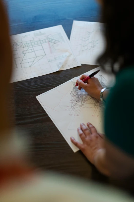 a woman sitting at a table writing on a piece of paper, a drawing, by Jessie Algie, unsplash, process art, architectural plans, peter eisenman, sustainable materials, patterns
