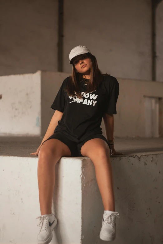 a woman sitting on top of a cement wall, by Adam Marczyński, wearing a black!! t - shirt, softglow, caps sideways, wearing shorts and t shirt