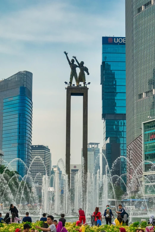 a group of people standing in front of a fountain, a statue, inspired by Cheng Jiasui, happening, jakarta, looming over a city, day time, raising between the buildings