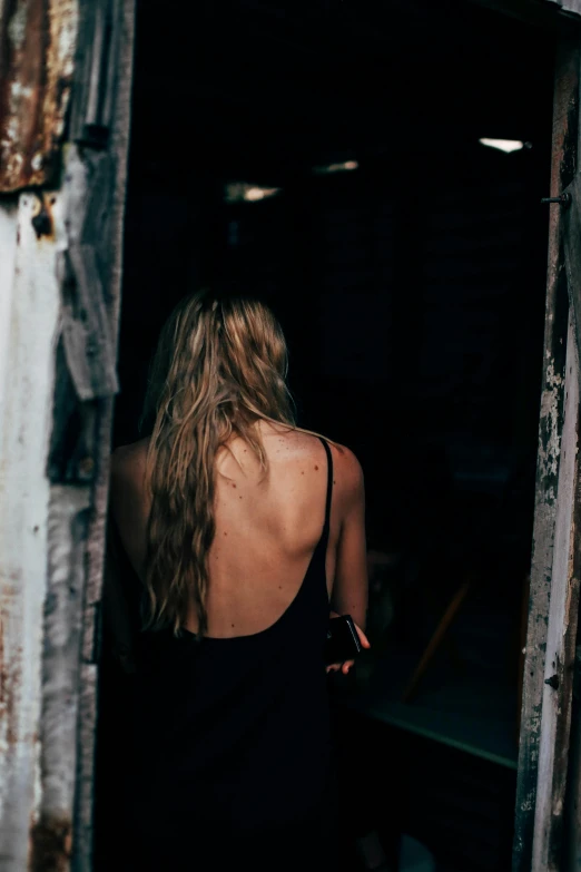 a woman in a black dress standing in a doorway, a picture, inspired by Elsa Bleda, unsplash contest winner, happening, messy blond hair, bare back, wearing a camisole, grungy