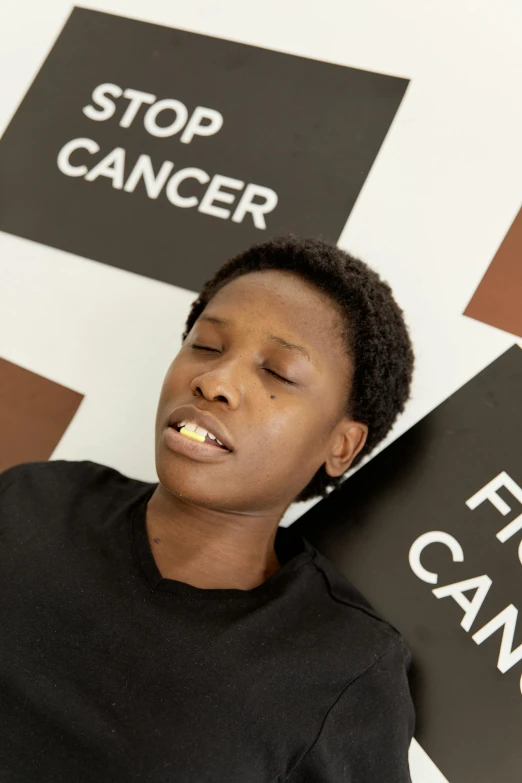 a woman holding a sign that says stop cancer, an album cover, chewing tobacco, emmanuel shiru, she is laying on her back, with a sad expression