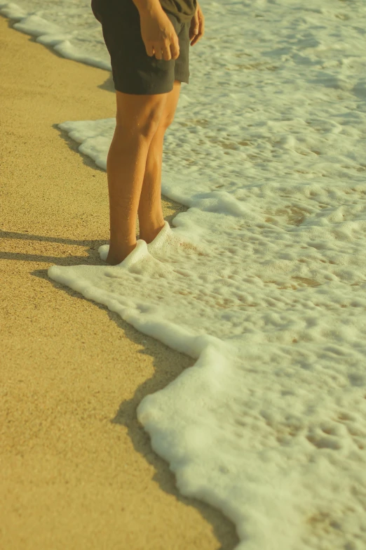 a person standing on a beach next to the ocean, sea foam, crooked legs, upclose, sunfaded