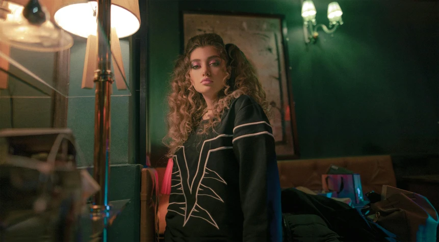 a woman standing in a room next to a lamp, an album cover, inspired by Elsa Bleda, trending on pexels, renaissance, in a black hoodie, her face framed with curls, still from a music video, greek ameera al taweel