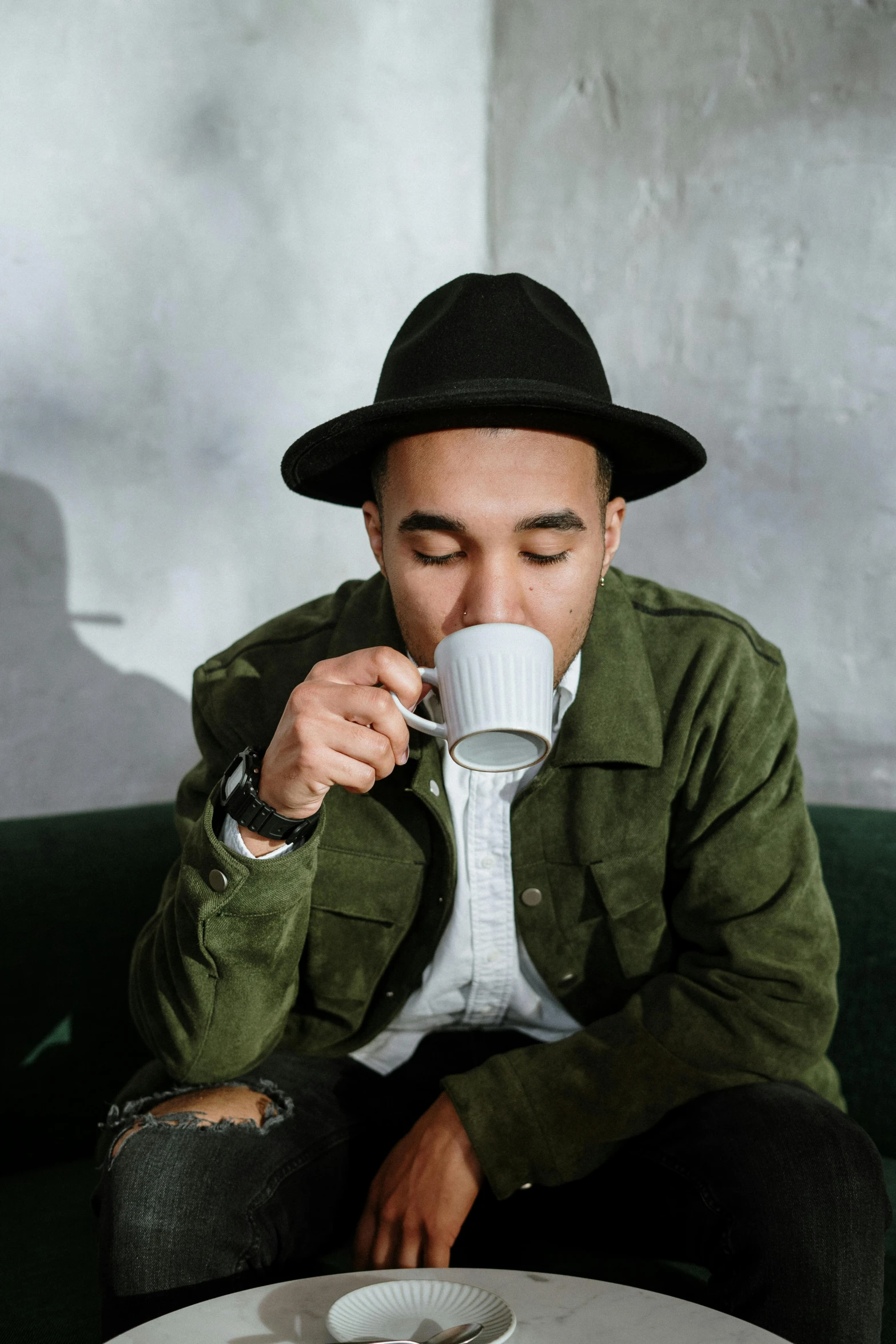 a man sitting on a couch drinking a cup of coffee, inspired by Michael Ray Charles, pexels contest winner, minimalism, wearing hat, mixed race, robert sheehan, tea cup