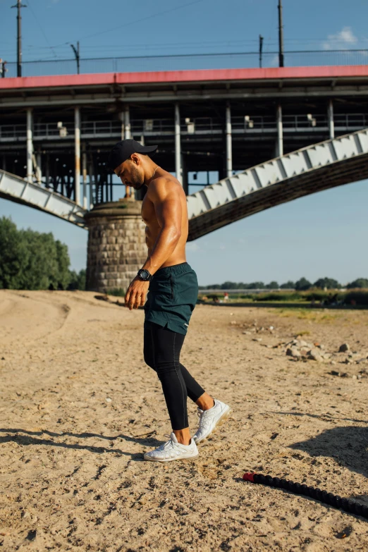 a man standing on top of a sandy beach next to a bridge, by Jan Tengnagel, unsplash, renaissance, attractive sporty physique, green legs, the thames is dry, full body potrait holding bottle