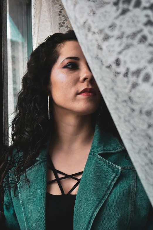 a woman in a green jacket looking out of a window, an album cover, inspired by Elsa Bleda, trending on pexels, renaissance, curly black hair, alanis guillen, wearing leather jacket, casual pose