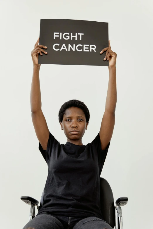 a woman sitting in a chair holding up a sign, by Ellen Gallagher, pexels contest winner, black arts movement, the cure for cancer, a person standing in front of a, fight pose, victor ngai