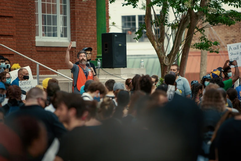 a man holding a microphone in front of a crowd of people, by Carey Morris, vandalism, square, tyler, exterior shot