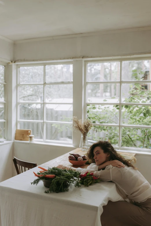 a woman sitting at a table in front of a window, inspired by Elsa Bleda, vegetables on table and candle, sleeping, in a white room, cottagecore