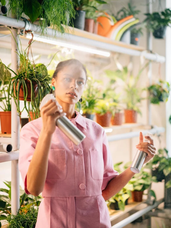 a woman standing in front of a shelf filled with potted plants, by Julia Pishtar, spraying liquid, profile image, clean lighting, flasks in hands