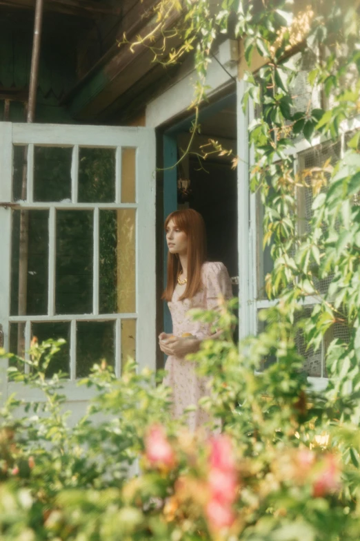 a woman in a pink dress looking out of a window, an album cover, unsplash, pre-raphaelitism, in a garden of a house, 70mm film screenshot, 4 k film still, amy pond