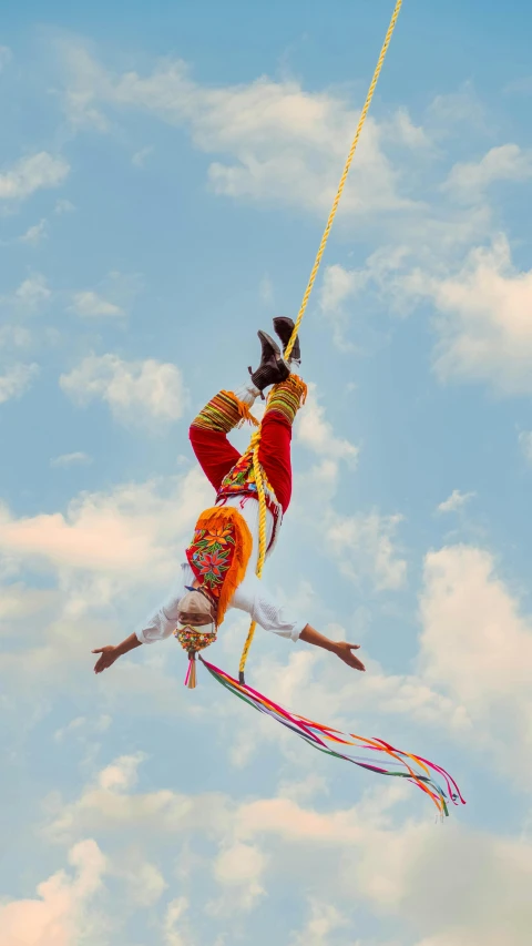 a person flying a kite high in the air, an album cover, pexels contest winner, arabesque, orange and yellow costume, ropes, folklorico, on stilts