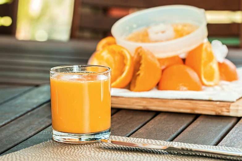 a glass of orange juice sitting on top of a wooden table, by Nicolette Macnamara, pexels, renaissance, 🦩🪐🐞👩🏻🦳, eating outside, grey orange, knockout punch