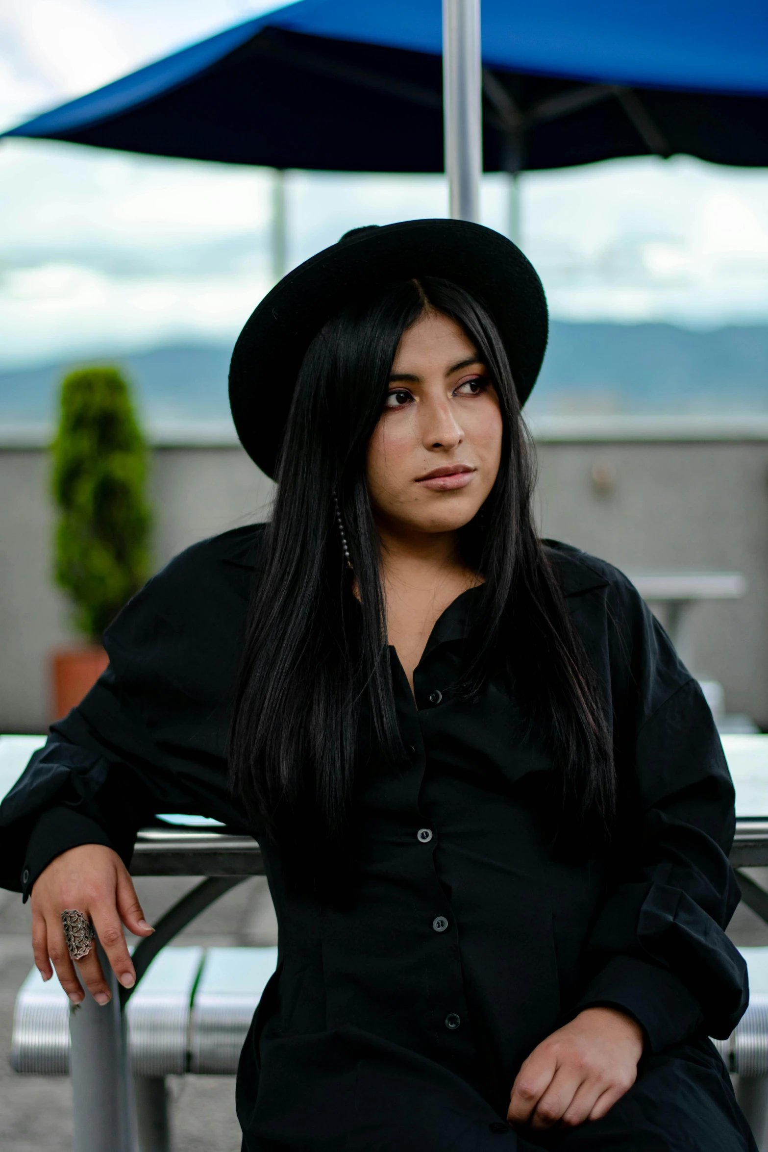 a woman sitting at a table with an umbrella in the background, wearing a black shirt, dark hat, latinas, profile image