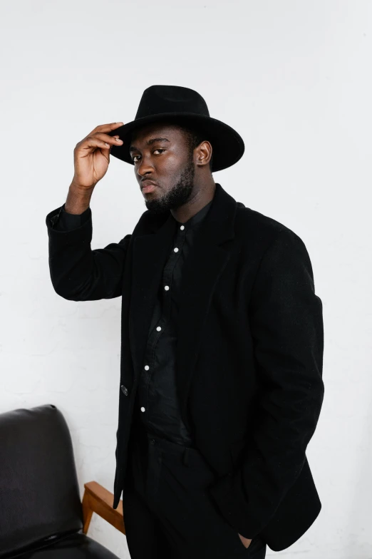 a man in a suit and hat standing next to a chair, inspired by Theo Constanté, pexels contest winner, jaylen brown, he is wearing a black trenchcoat, slightly minimal, black skin!!!