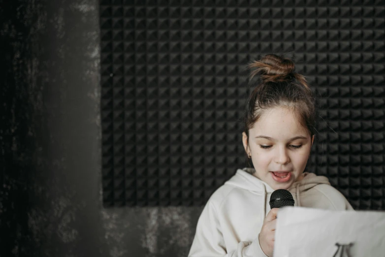 a girl reading a piece of paper in front of a microphone, an album cover, by Emma Andijewska, trending on pexels, kid, background image, closed mouth, in small room