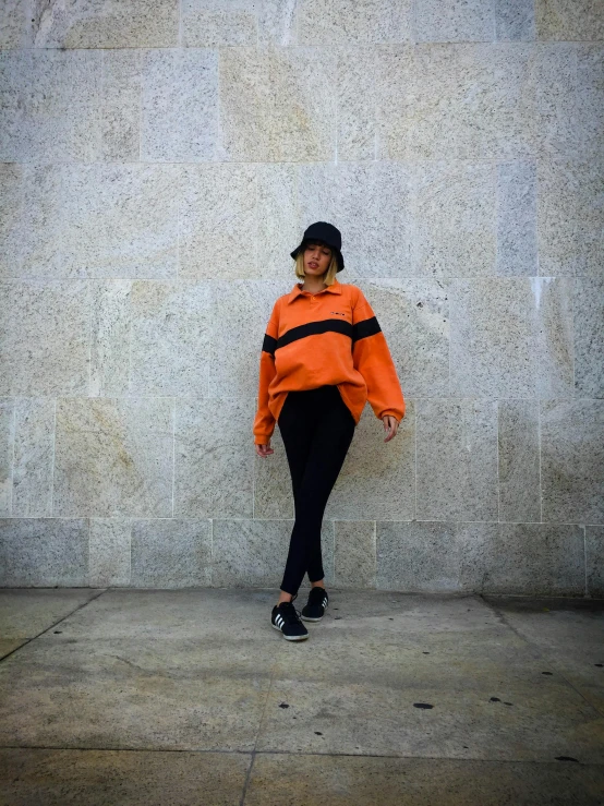 a woman standing in front of a stone wall, an album cover, by Robbie Trevino, trending on unsplash, long orange sweatshirt, wearing track and field suit, orange and black, full body profile camera shot