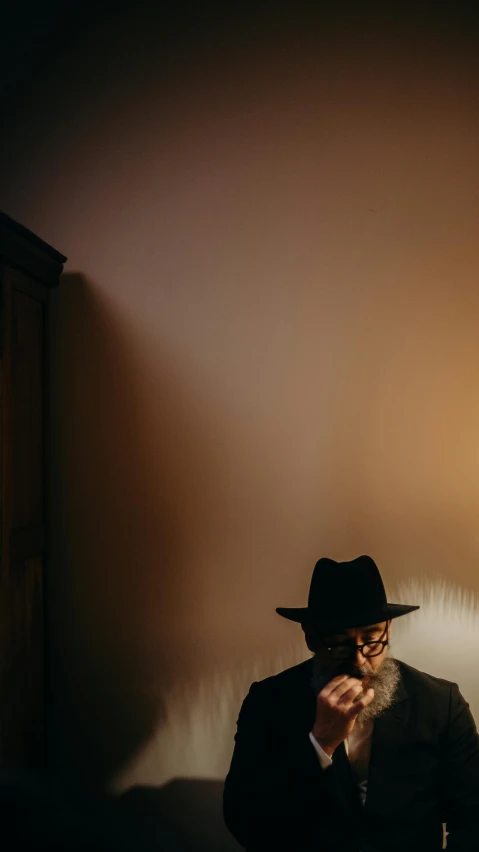 a man sitting on top of a bed next to a lamp, an album cover, pexels, dark hat, cowboy hat and glasses, standing in a dimly lit room, profile shot