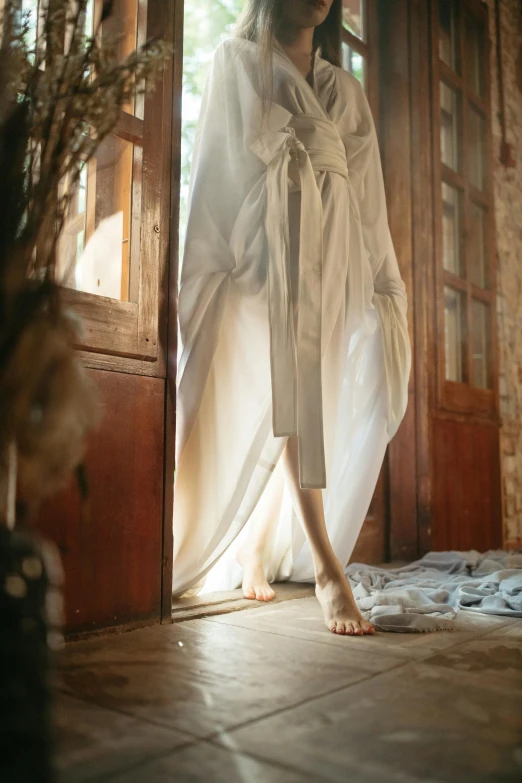 a woman in a white robe standing in a doorway, a picture, inspired by Anka Zhuravleva, unsplash contest winner, renaissance, draped in flowing fabric, legs visible, indoors, silver，ivory