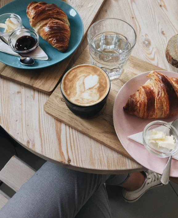 a person sitting at a table with a plate of croissants and a cup of coffee, by Alice Mason, trending on unsplash, thumbnail, restaurant menu photo, a wooden, taken from the high street