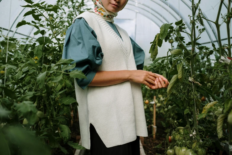 a woman standing in a greenhouse with tomatoes, an album cover, inspired by Modest Urgell, unsplash, hurufiyya, model is wearing techtical vest, wearing a white sweater, teal tunic, thumbnail
