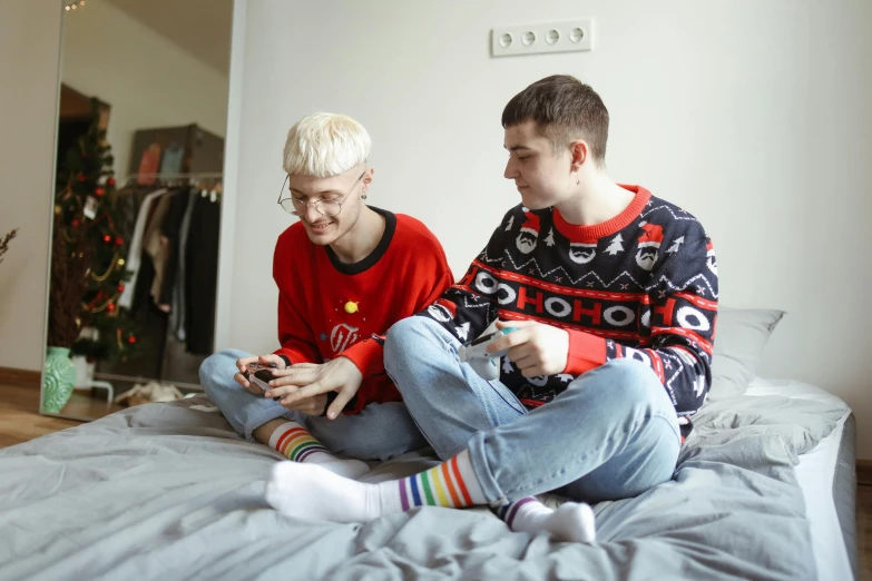 a couple of young men sitting on top of a bed, a photo, trending on pexels, pixel art, wearing festive clothing, genderless, playing games, red sweater and gray pants