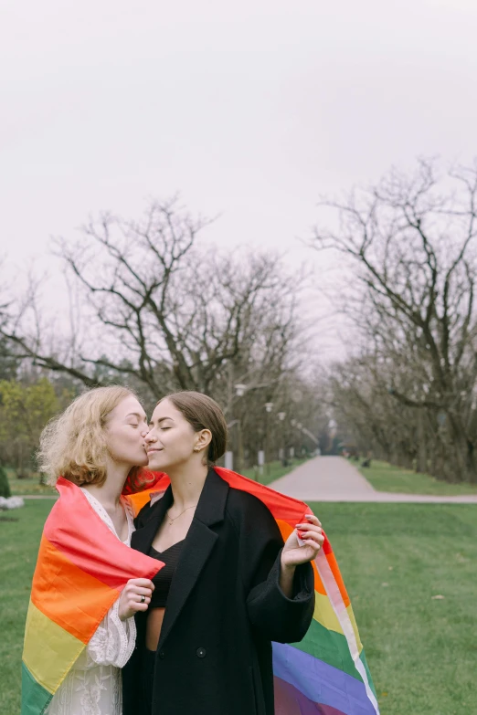 a couple of women standing on top of a lush green field, by Attila Meszlenyi, trending on unsplash, romanticism, black and orange coat, two women kissing at a carnival, in a park, lgbt flag